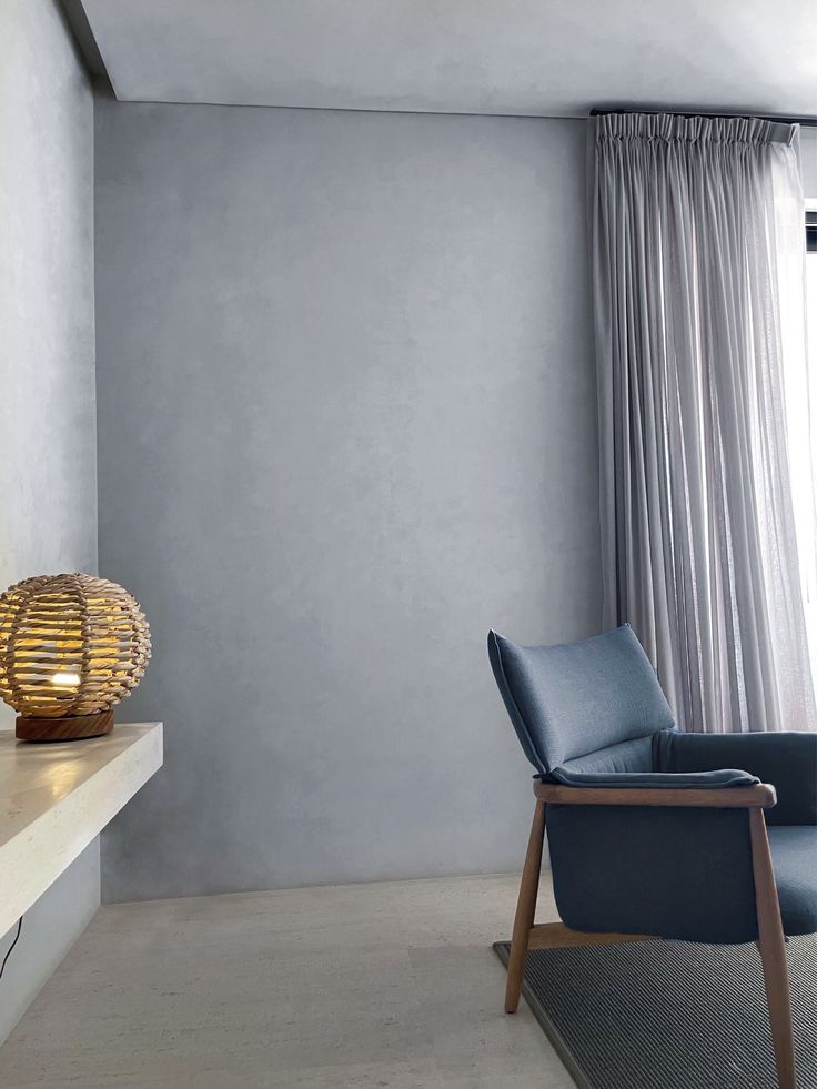 a blue chair sitting in front of a window next to a wooden table with a lamp on it