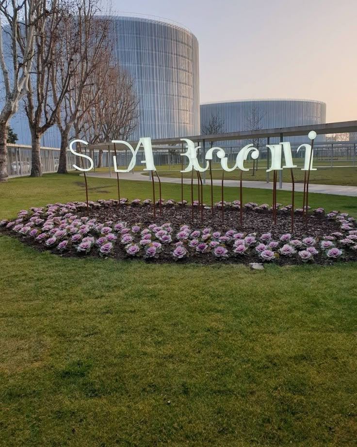 a sign that is in the middle of some grass with flowers around it and trees