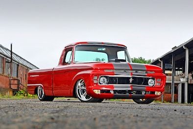 an old red pickup truck parked in front of a building with two barns behind it