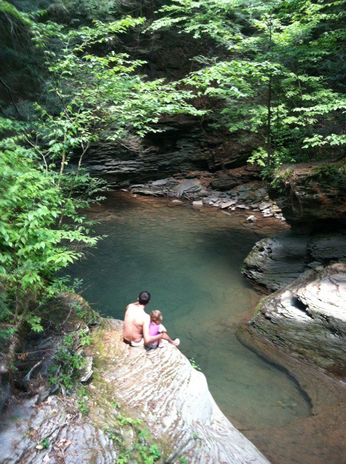 two people are sitting on the edge of a cliff next to a river and some trees