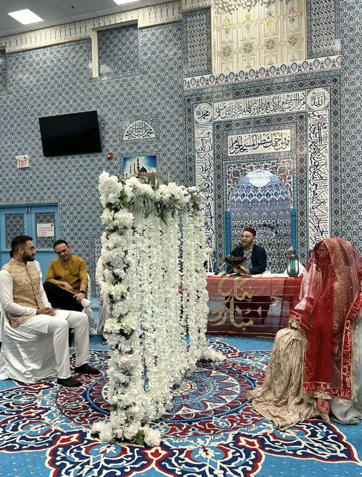 a group of people sitting in chairs around a table with flowers on it and two men standing next to each other