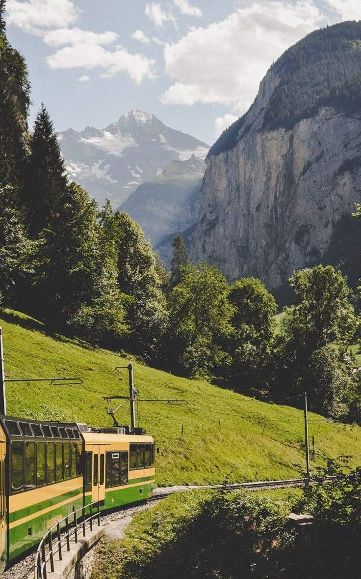 a green and yellow train traveling through a lush green forest filled mountain side covered in snow capped mountains