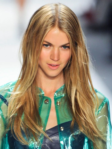 a model walks down the runway in a shiny green jacket and blue skirt with gold buttons