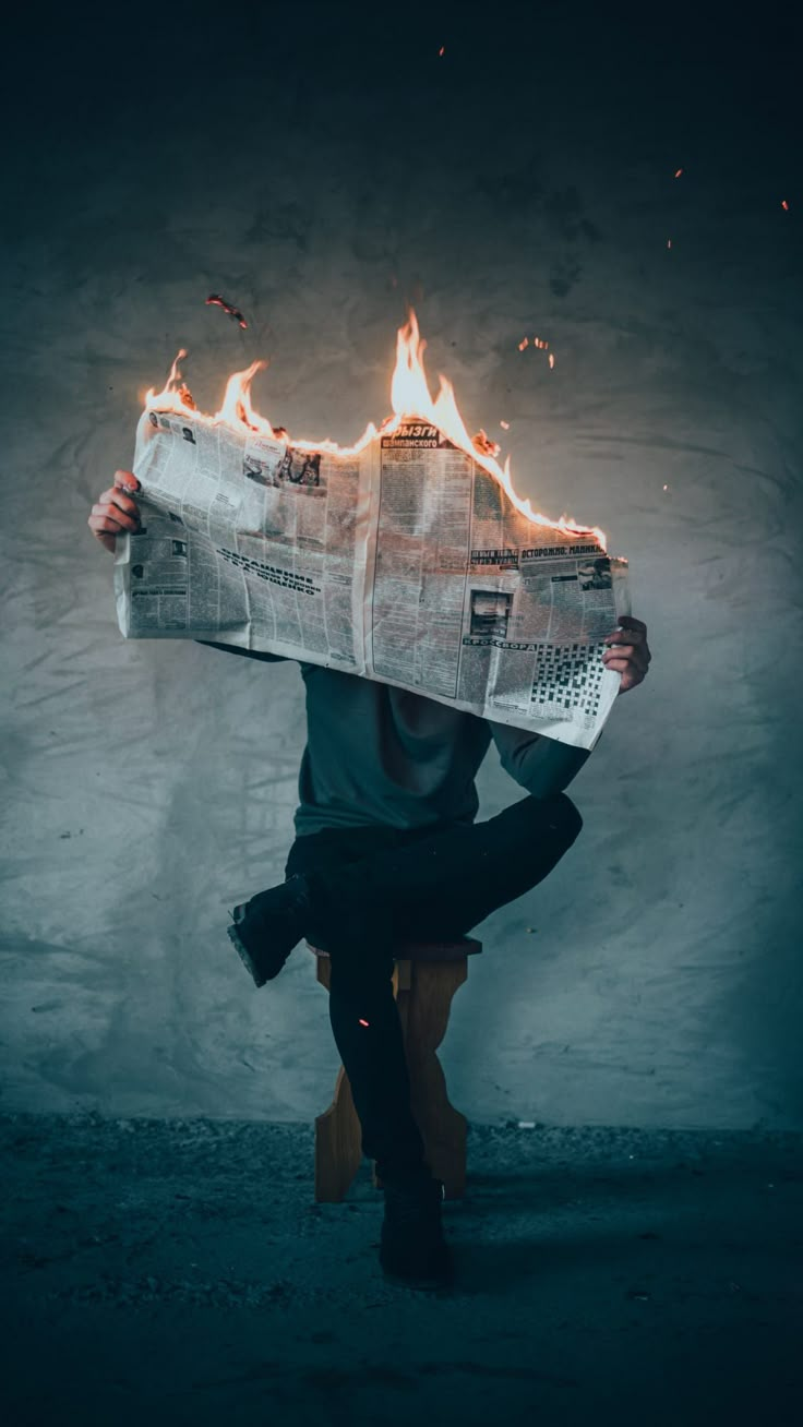 a man sitting on a chair holding a newspaper with flames coming out of it and reading the paper