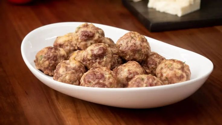 a white bowl filled with meatballs sitting on top of a wooden table next to cheese