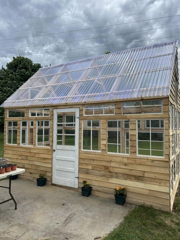 a small wooden house with a metal roof and glass windows on the side, surrounded by potted plants