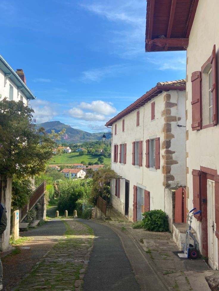 a narrow street with red shutters on both sides and green hills in the distance