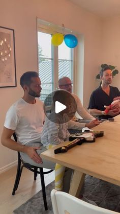 three men sitting at a table talking to each other in a room with balloons on the wall