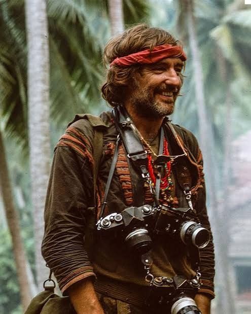 a man with a camera on his shoulder walking through the woods in front of palm trees