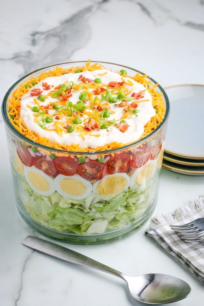 the 7 layer salad is in a glass bowl with ingredients labeled below it and on top of each serving dish