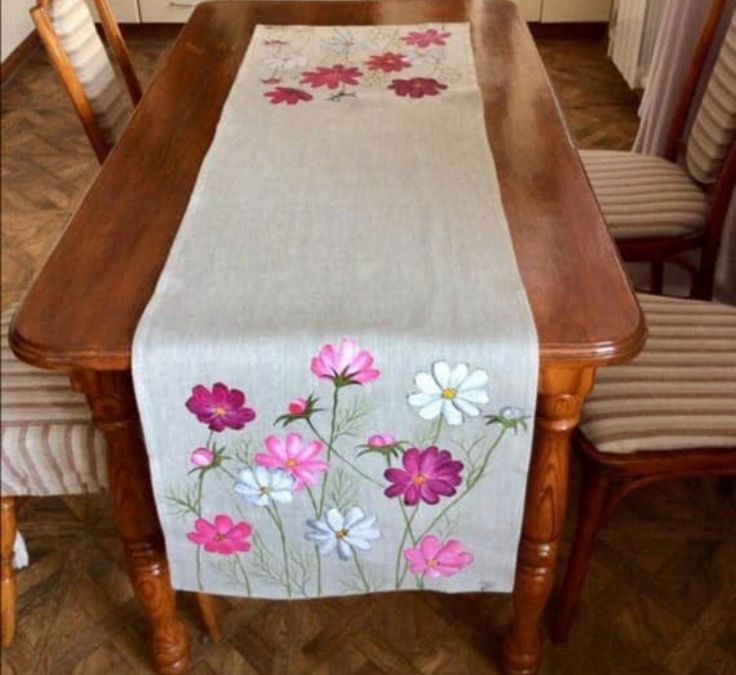 a dining room table with flowers painted on the table runner and chairs around it, in front of a kitchen window