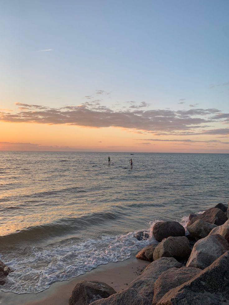 two people in the ocean at sunset with rocks on the shore and one person walking out into the water