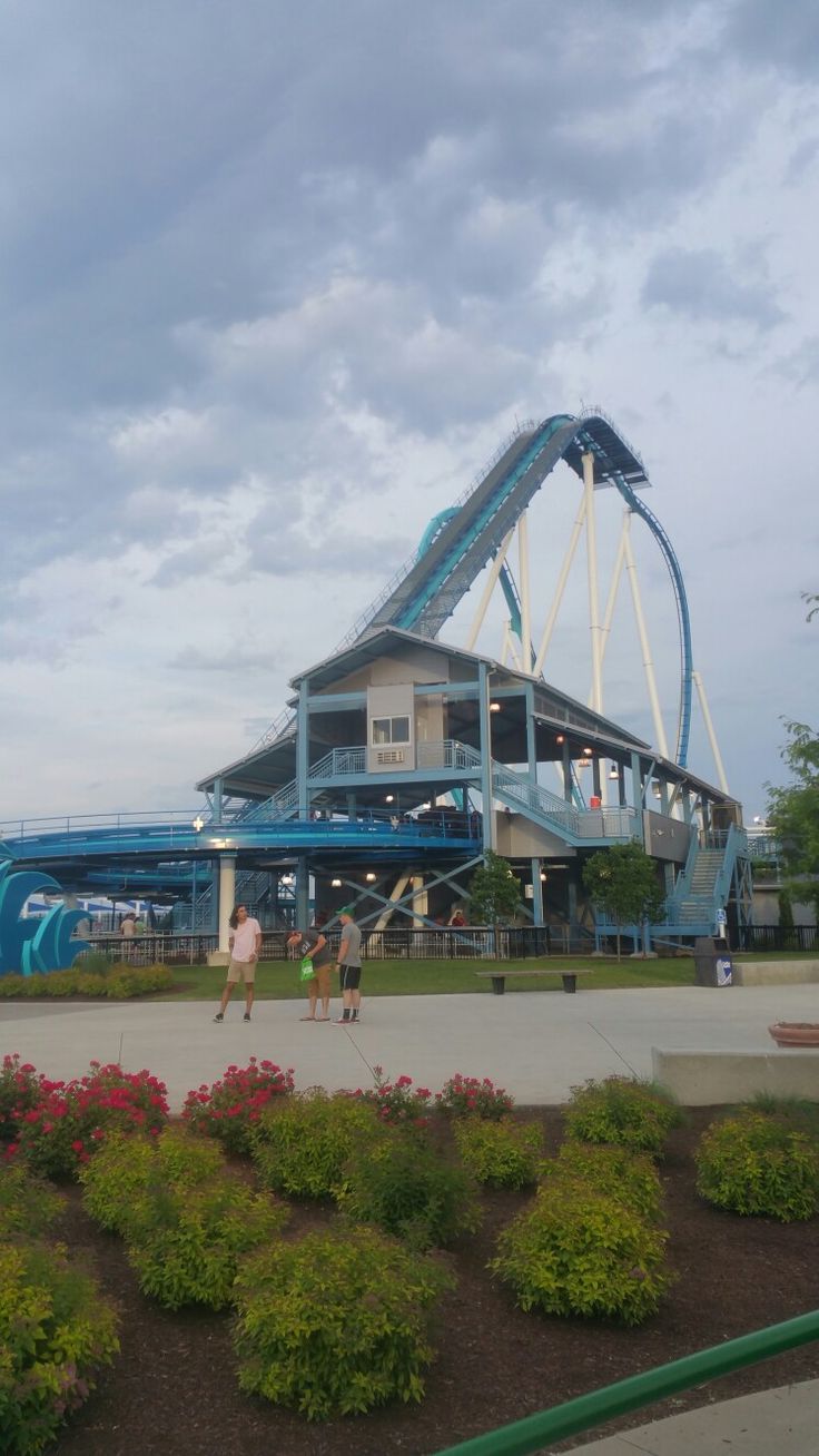 people are walking around in front of a roller coaster at the amusement park on a cloudy day