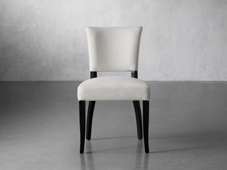 a white chair with black legs in front of a gray wall and concrete flooring