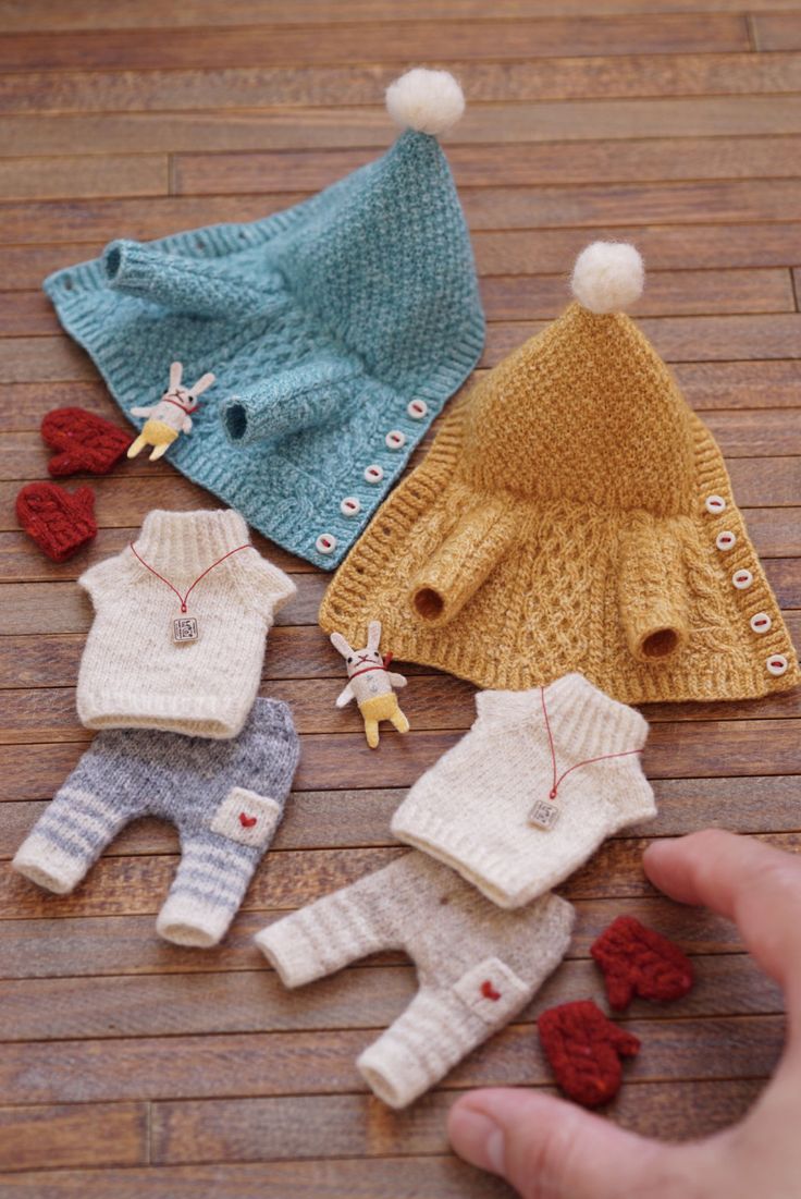 four knitted baby sweaters and pants on a wooden floor, one being held up by a person's hand