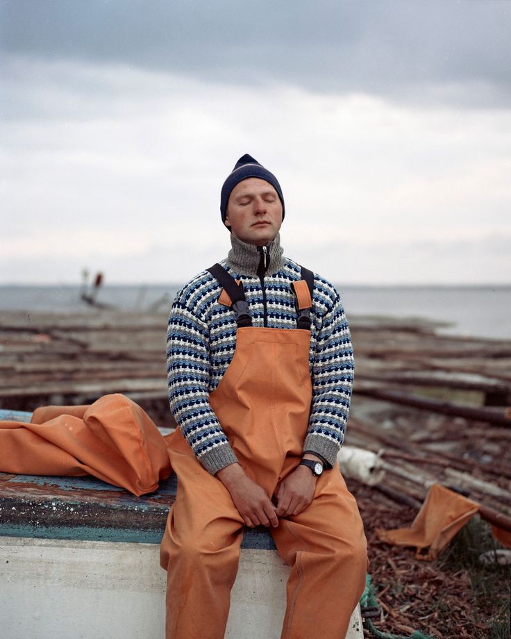 a man wearing an orange bib and overalls sitting on a wall next to the ocean