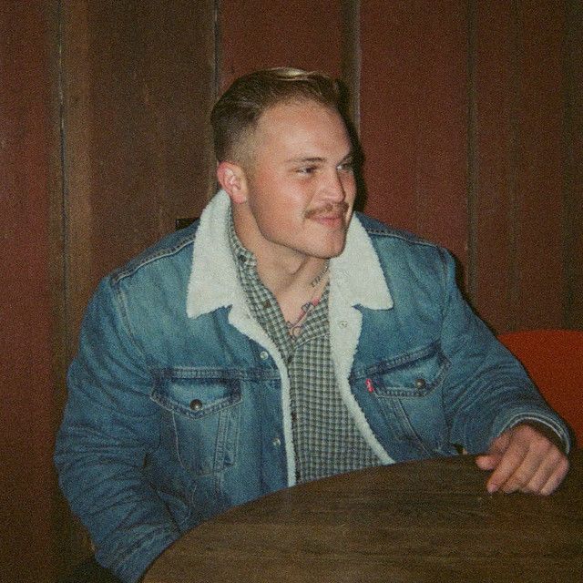 a man sitting at a table in front of a wooden paneled wall and wearing a denim jacket