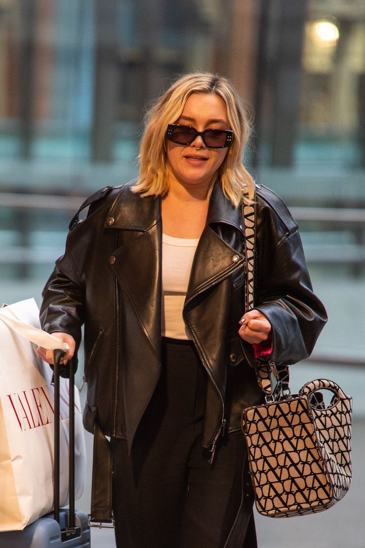 a woman in black leather jacket and sunglasses with luggage walking down the street while holding her handbag