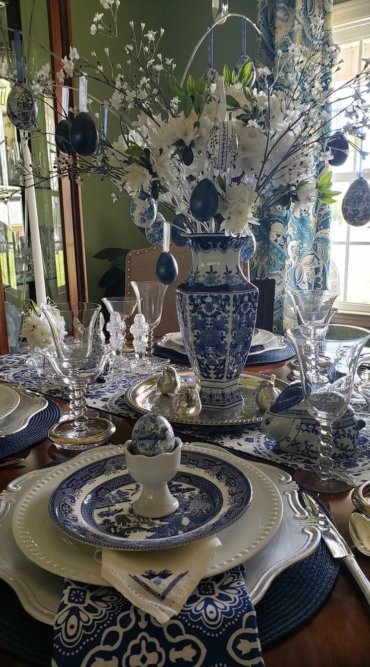 a dining room table set with blue and white plates, silverware and vases