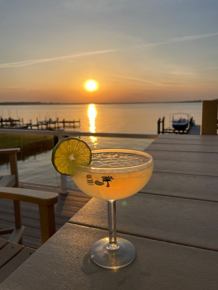 a margarita sitting on top of a wooden table next to the ocean at sunset with a slice of lemon