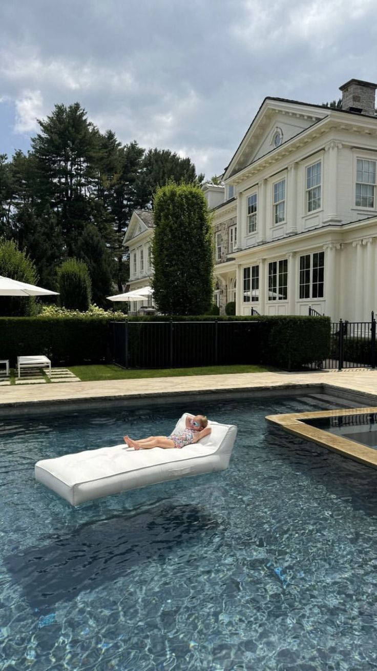 a woman laying on an inflatable mattress next to a swimming pool with a house in the background