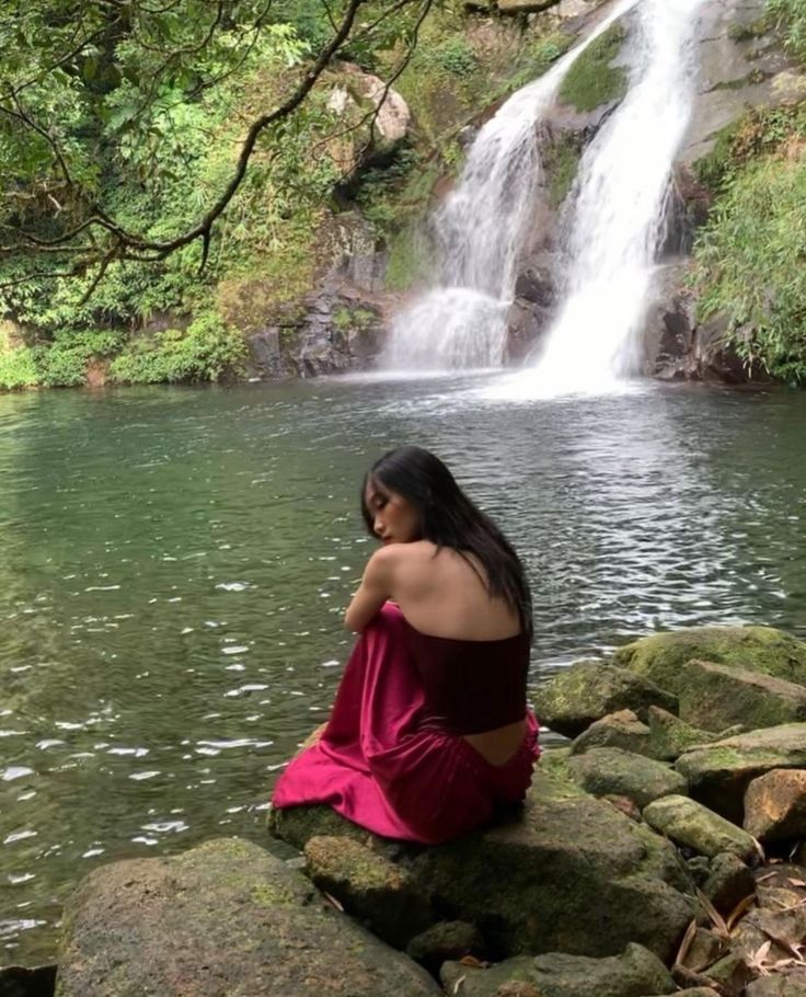 a woman in a red dress sitting on rocks near a river with a waterfall behind her