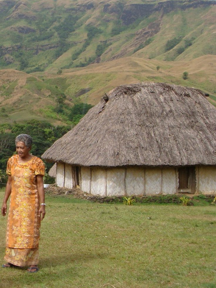 a woman standing in front of a thatched hut with mountains in the back ground