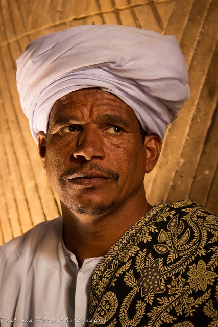 a man wearing a white turban standing in front of a bamboo wall and looking at the camera