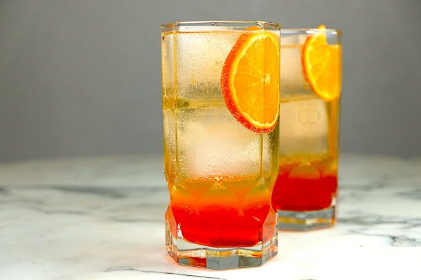 two glasses filled with orange and red liquid on top of a marble counter next to each other