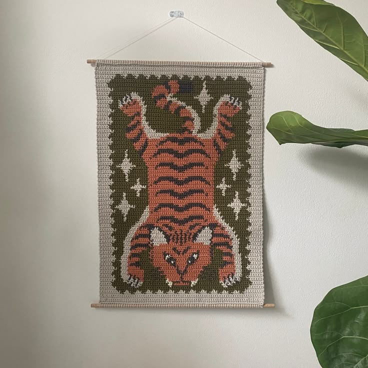 a cross - stitch tiger hanging on a wall next to a potted green plant