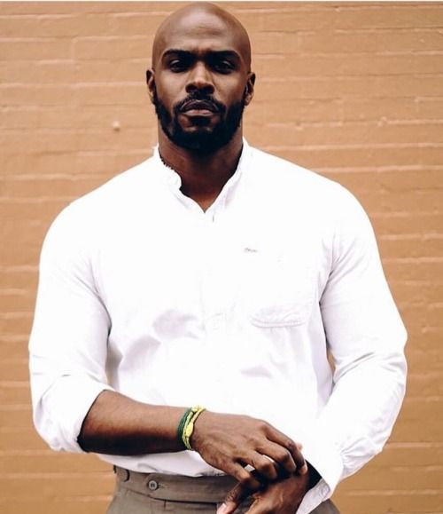 a man standing in front of a brick wall wearing a white shirt and khaki pants