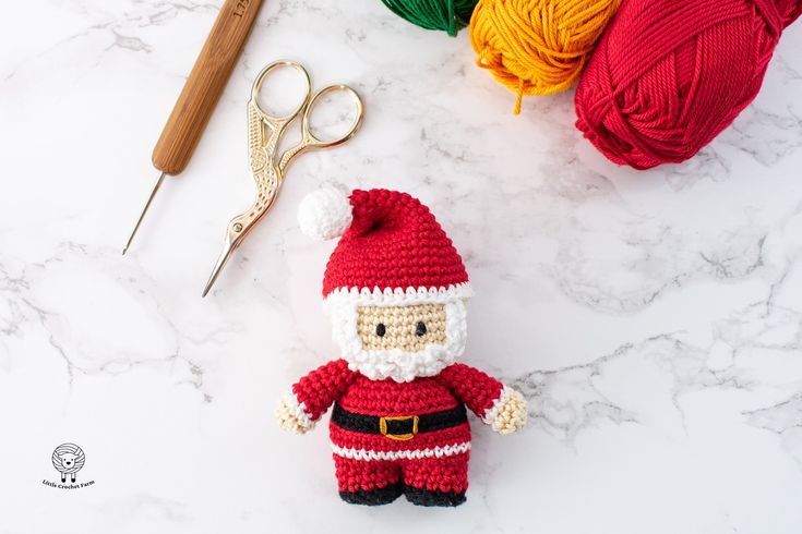 a crocheted santa clause doll next to yarn and scissors on a marble surface