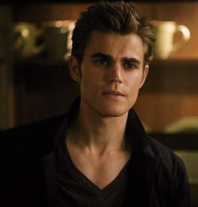 a young man with dark hair and black shirt looking at the camera in a dimly lit room