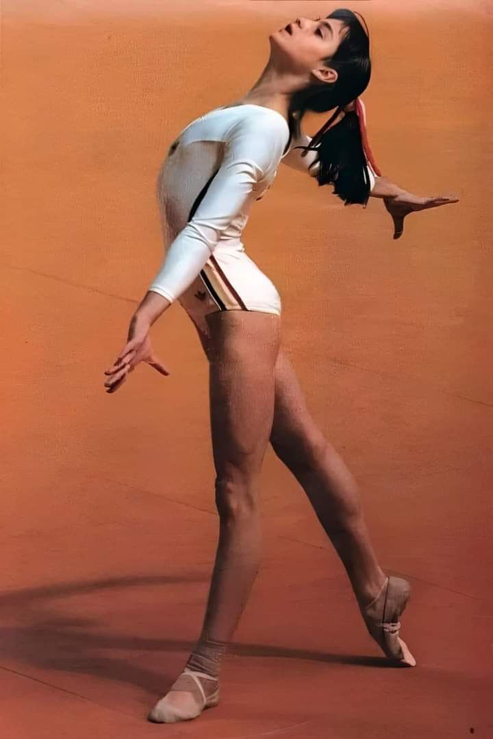 a woman in white leotard and ballet shoes standing on a tennis court with her arms stretched out