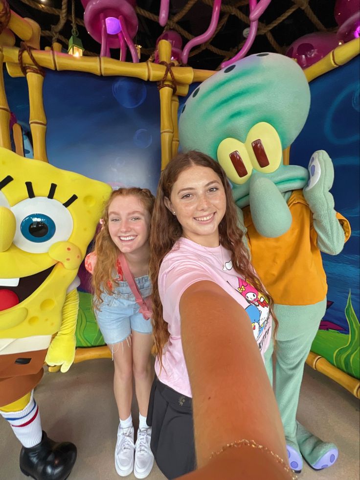 two girls standing in front of spongebob characters at an amusement park with their arms around each other