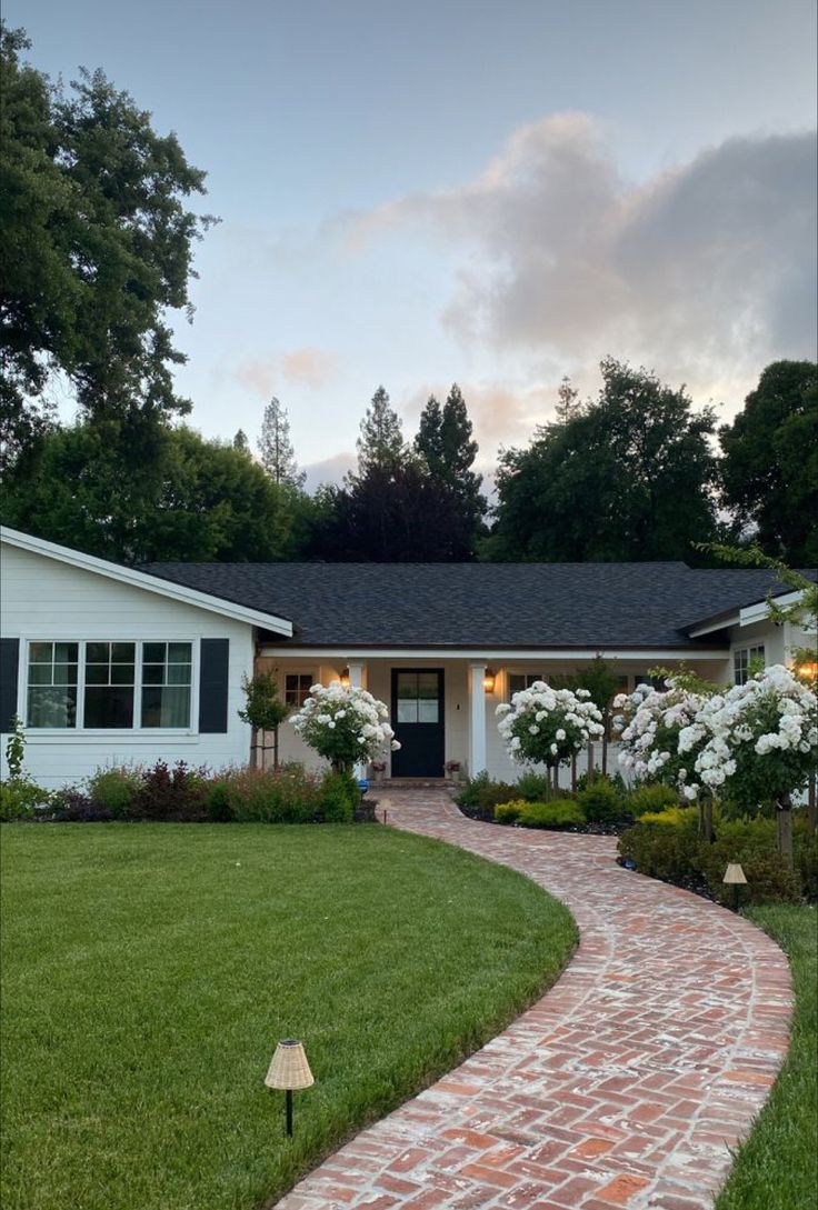 a brick path leading to a house with white flowers in the front yard and landscaping around it