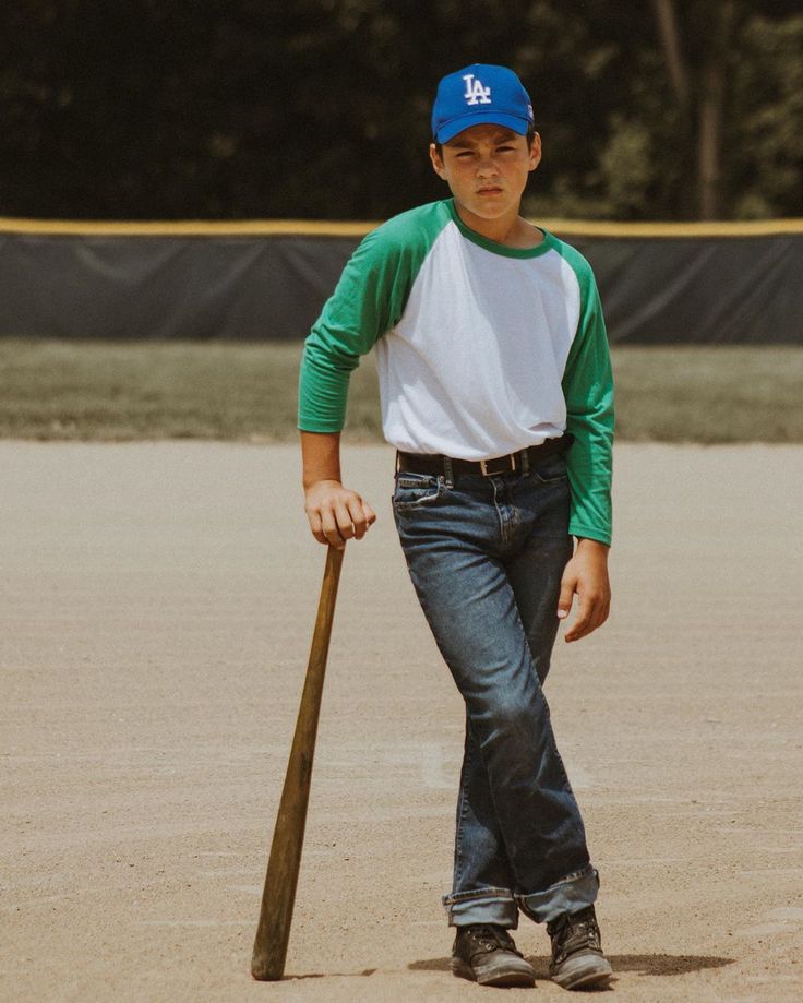 a young man holding a baseball bat on top of a dirt field in front of trees