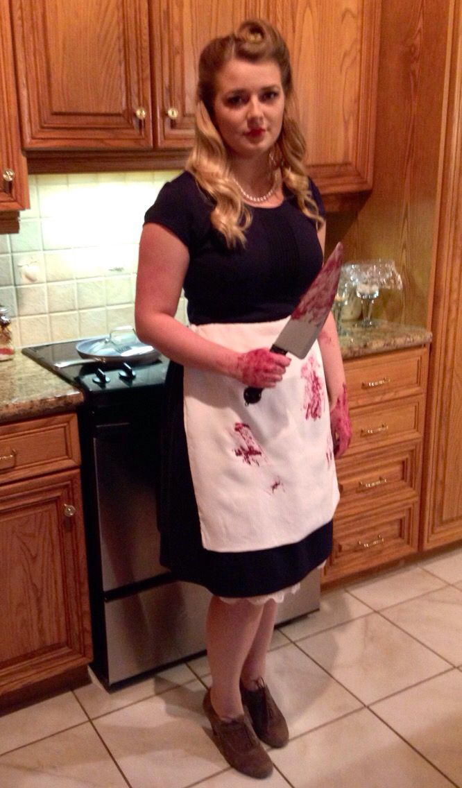 a woman standing in a kitchen holding a knife and wearing an apron with blood on it