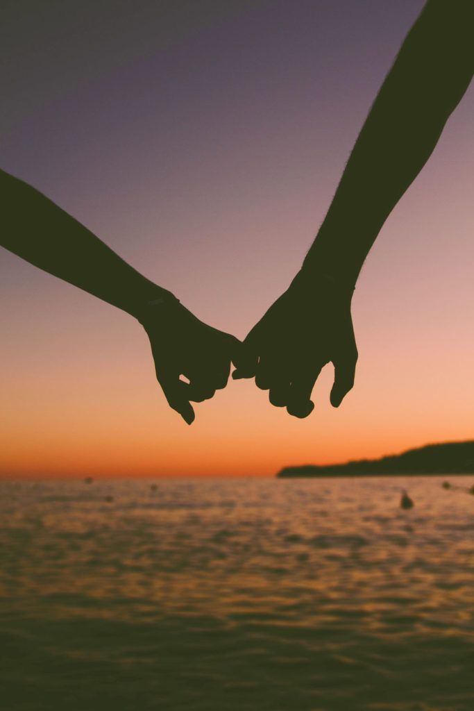 two people holding hands while standing in front of the ocean at sunset with boats in the background