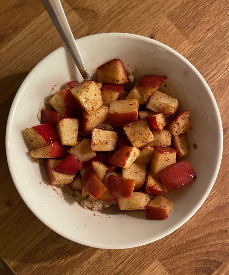 a white bowl filled with cooked apples on top of a wooden table next to a spoon