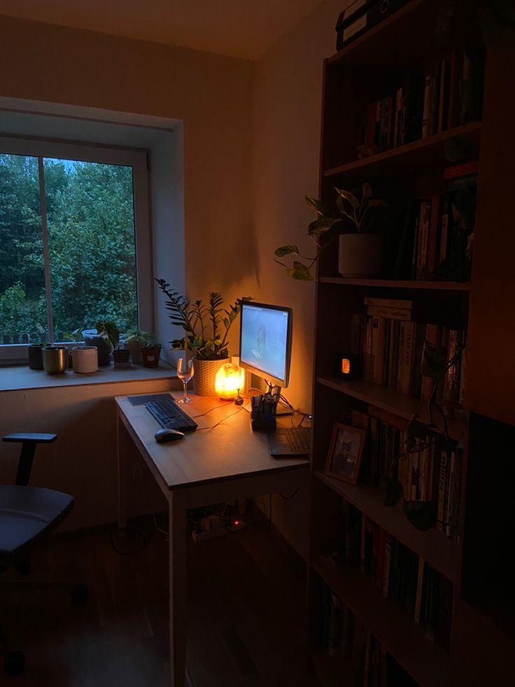 a dimly lit room with a computer and bookshelf in front of the window