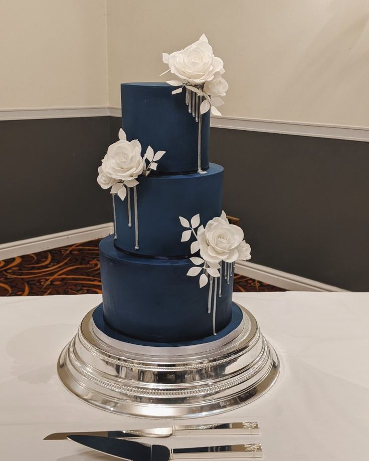 a three tiered blue cake with white flowers on the top and silver serving utensils