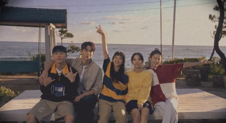 a group of people sitting on top of a wooden bench next to the ocean with their arms in the air