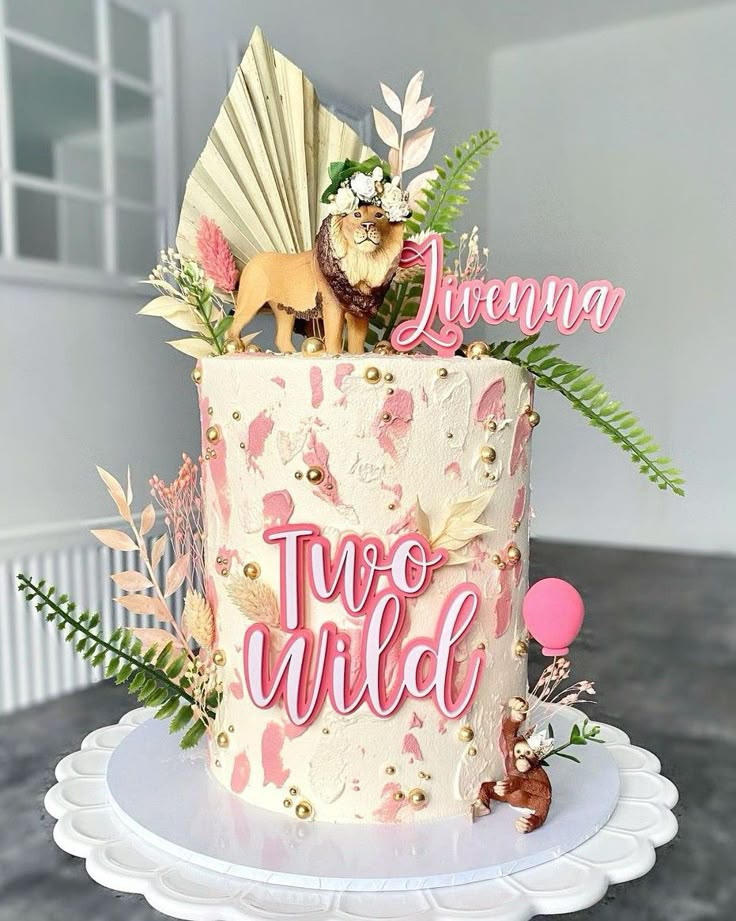 a pink and white cake with the words two wild on top, surrounded by tropical leaves