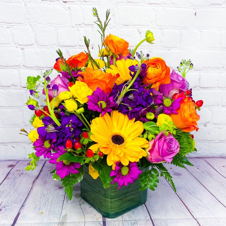 a square vase filled with colorful flowers on top of a wooden table next to a white brick wall