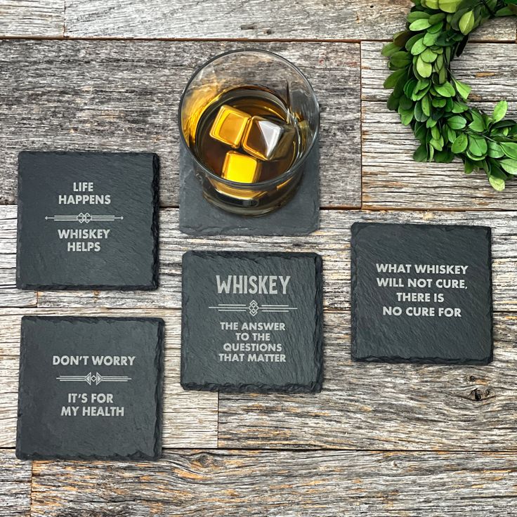 four slate coasters with sayings on them sitting next to a glass of whiskey