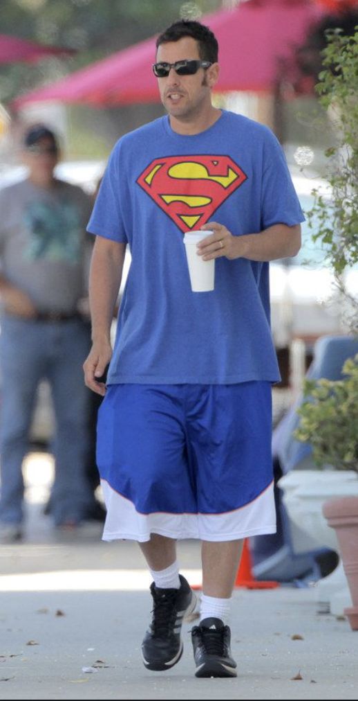 a man in a superman t - shirt and shorts walks down the street while holding a drink