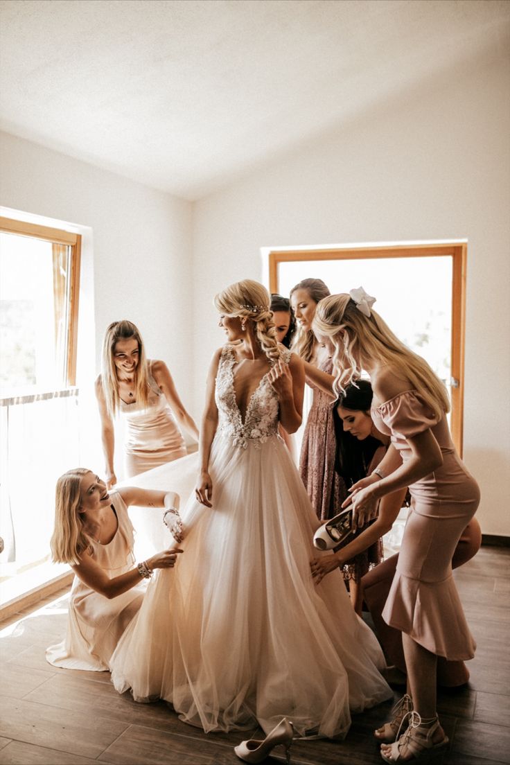 a group of women standing around each other in front of a window with one woman helping another woman put on her wedding dress