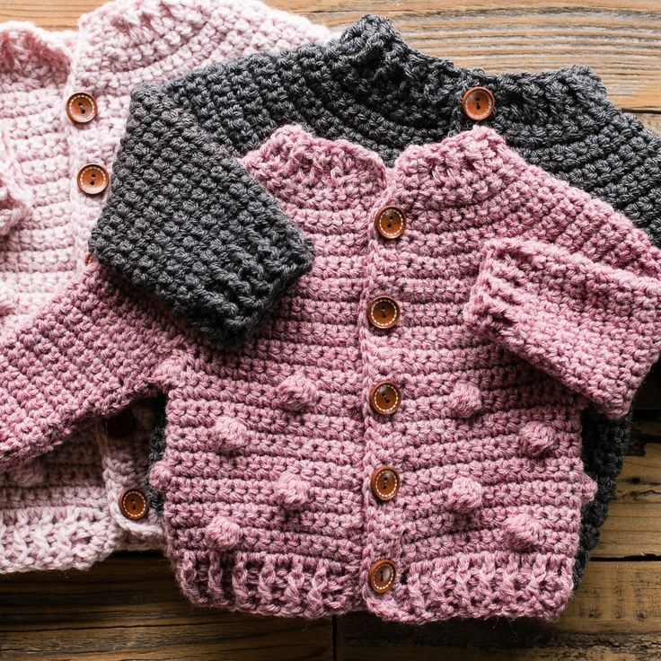 three crocheted sweaters are sitting on top of a wooden table, one is pink and the other is gray