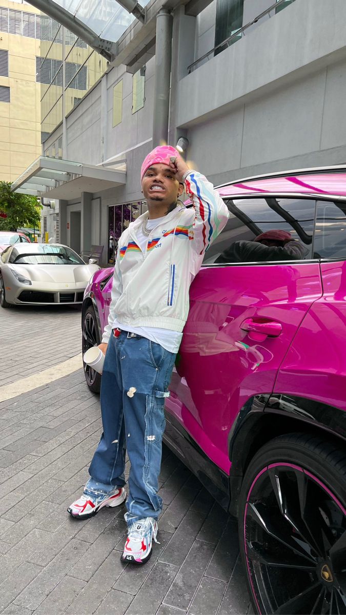 a young man leaning on the side of a pink car in front of a building